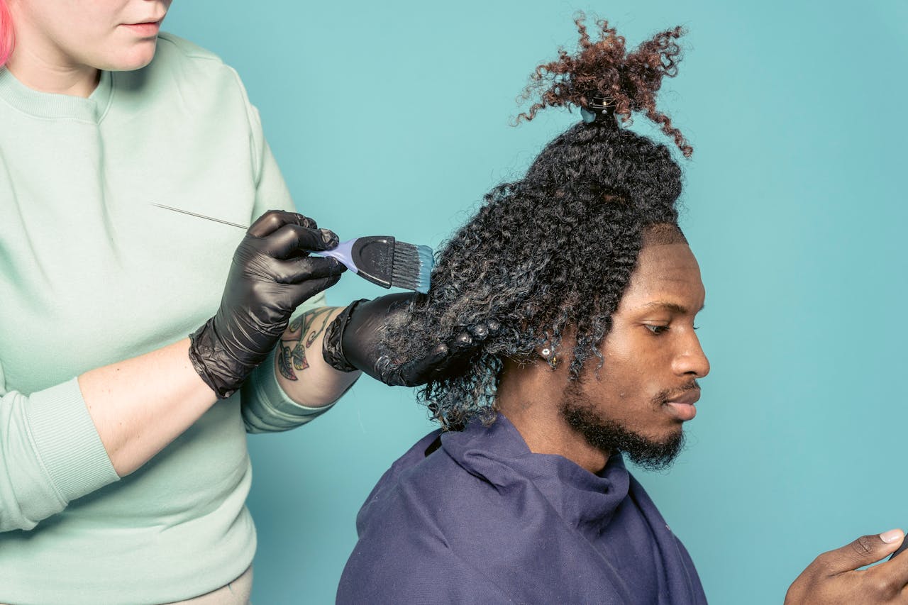 Services Crop hairstylist with tattoo dying African American male customer using cellphone against blue background