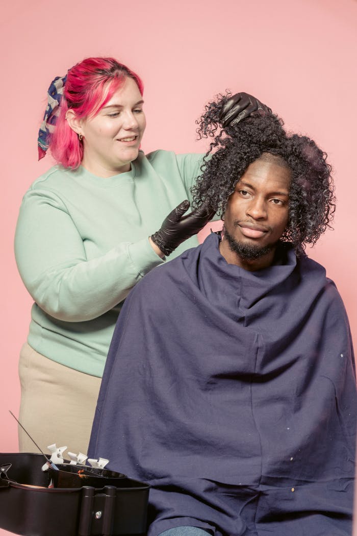 Home Female hairdresser in gloves standing behind African American customer and preparing hair for spreading dye while working in studio against pink background