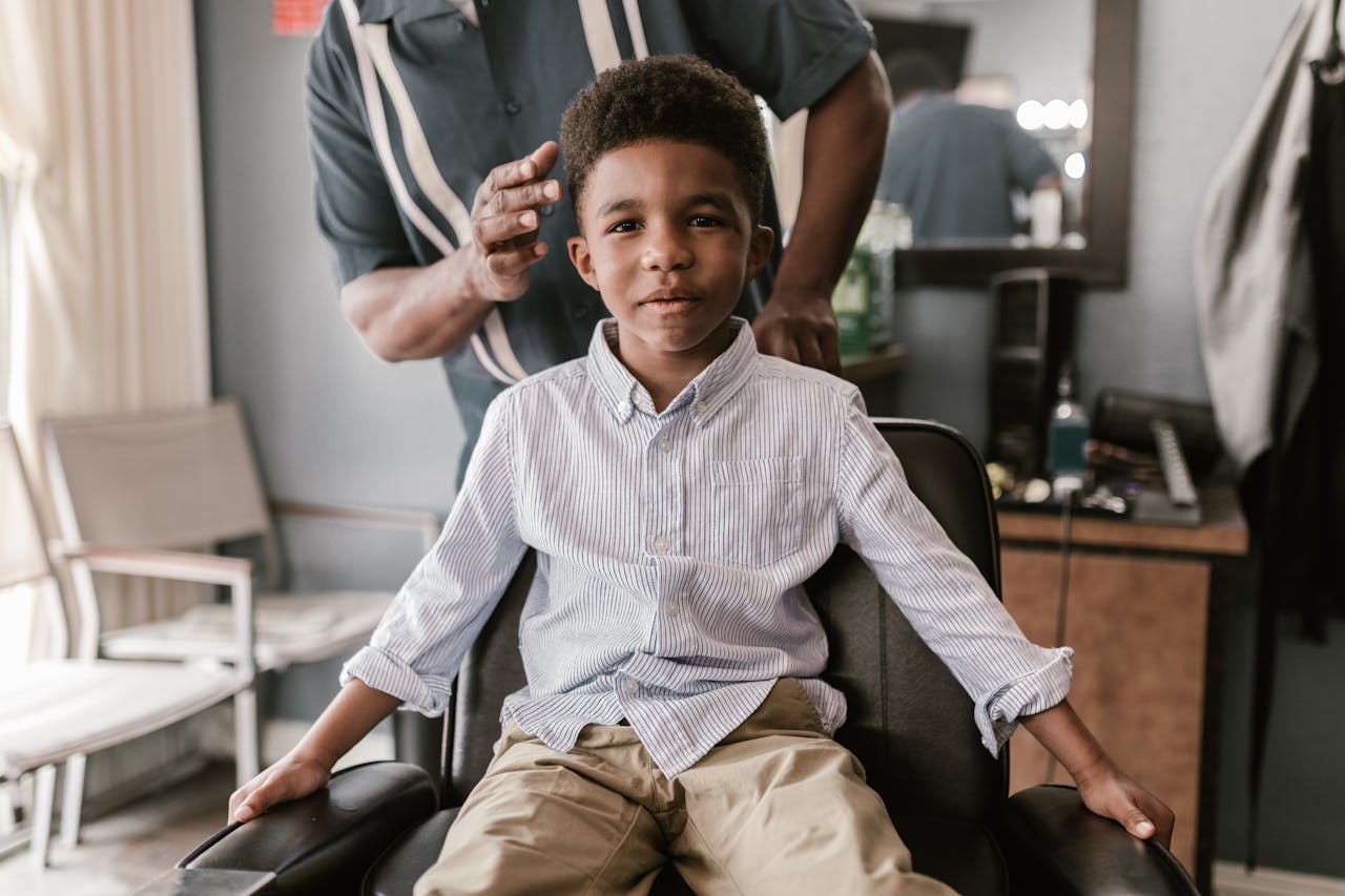 Home A young boy in a striped shirt smiles while seated in a barbershop chair, anticipating a haircut.