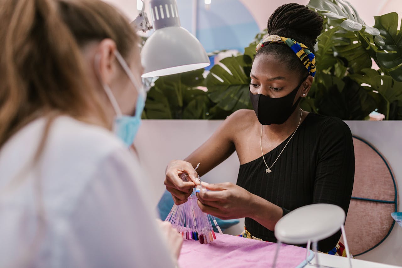 Services A nail technician and customer wearing masks in a modern salon choosing nail polish colors, embracing safety measures.